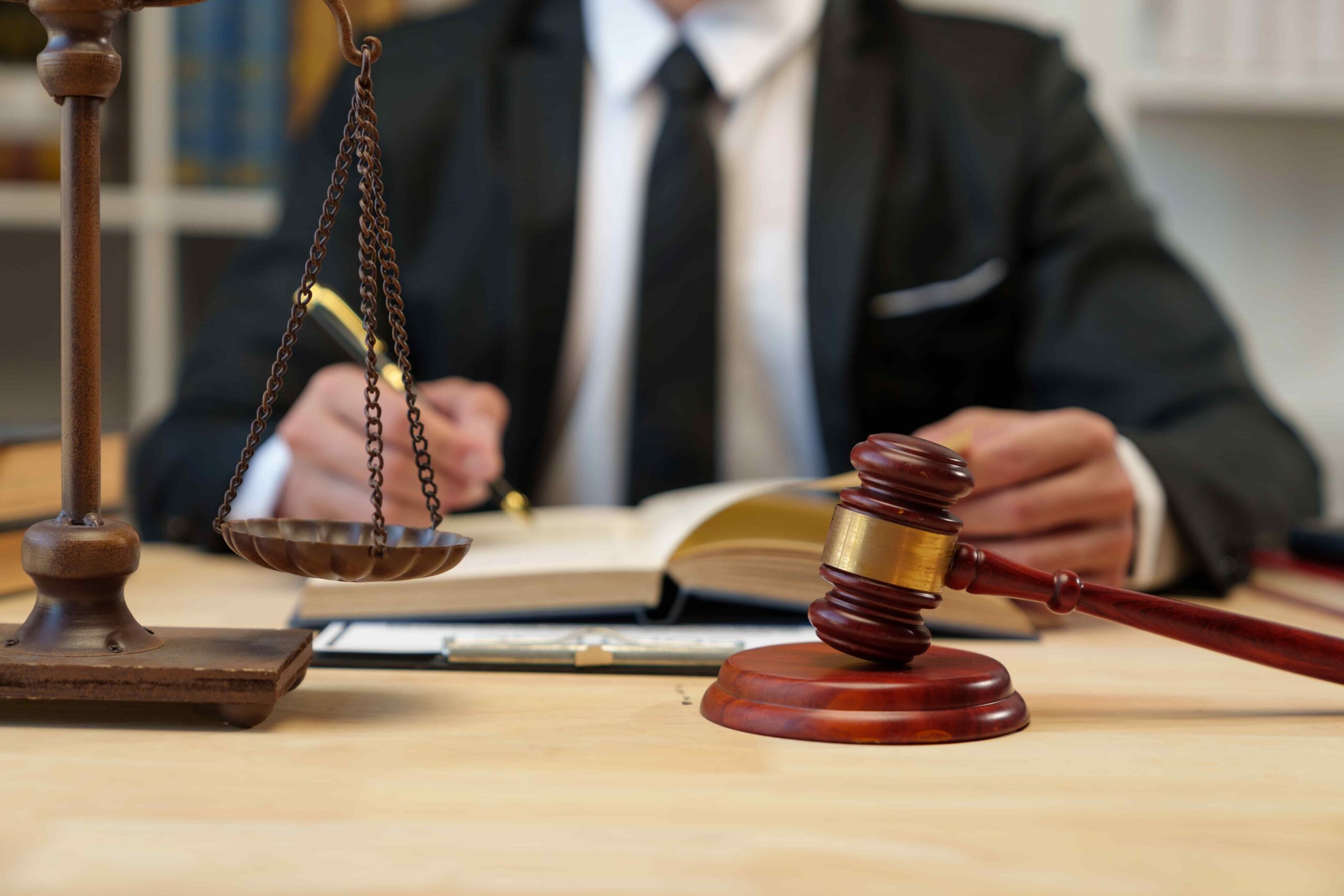 Judge’s gavel and scales of justice on a desk with a lawyer writing in a law book, symbolizing legal proceedings, justice, and courtroom decision-making.