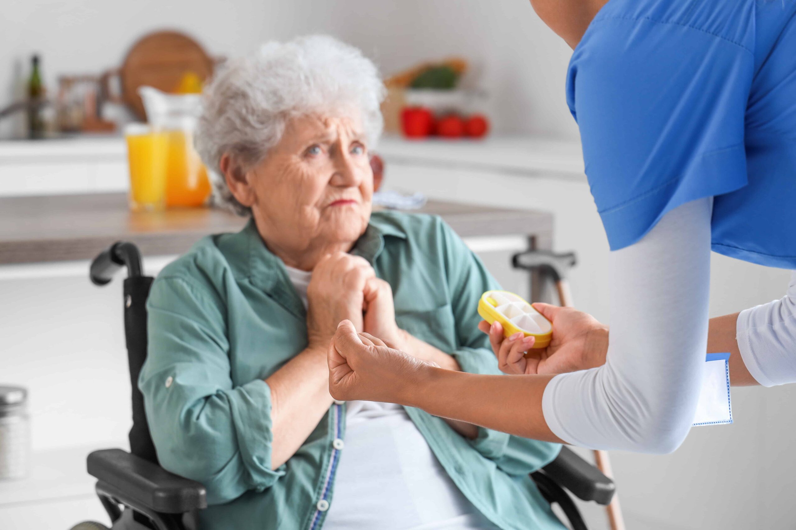 Elderly woman in a wheelchair receiving medication assistance from a caregiver at home.